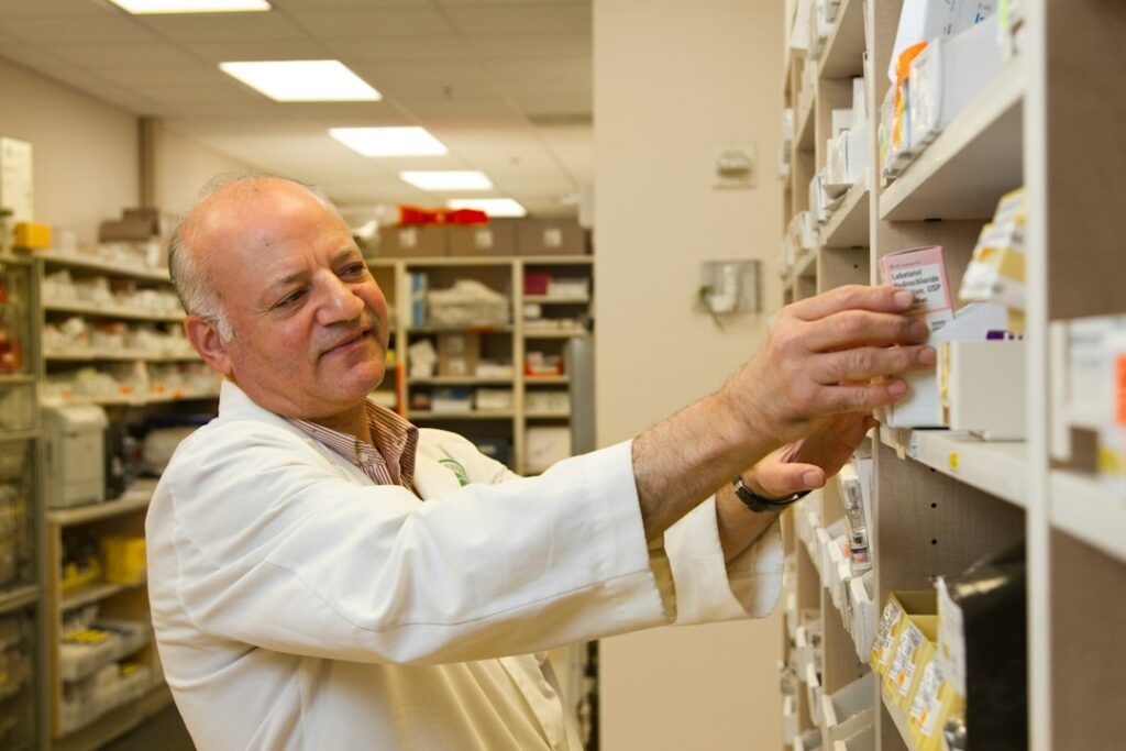 A person speaks with a pharmacist while holding a prescription bottle beside a pharmacy counter.